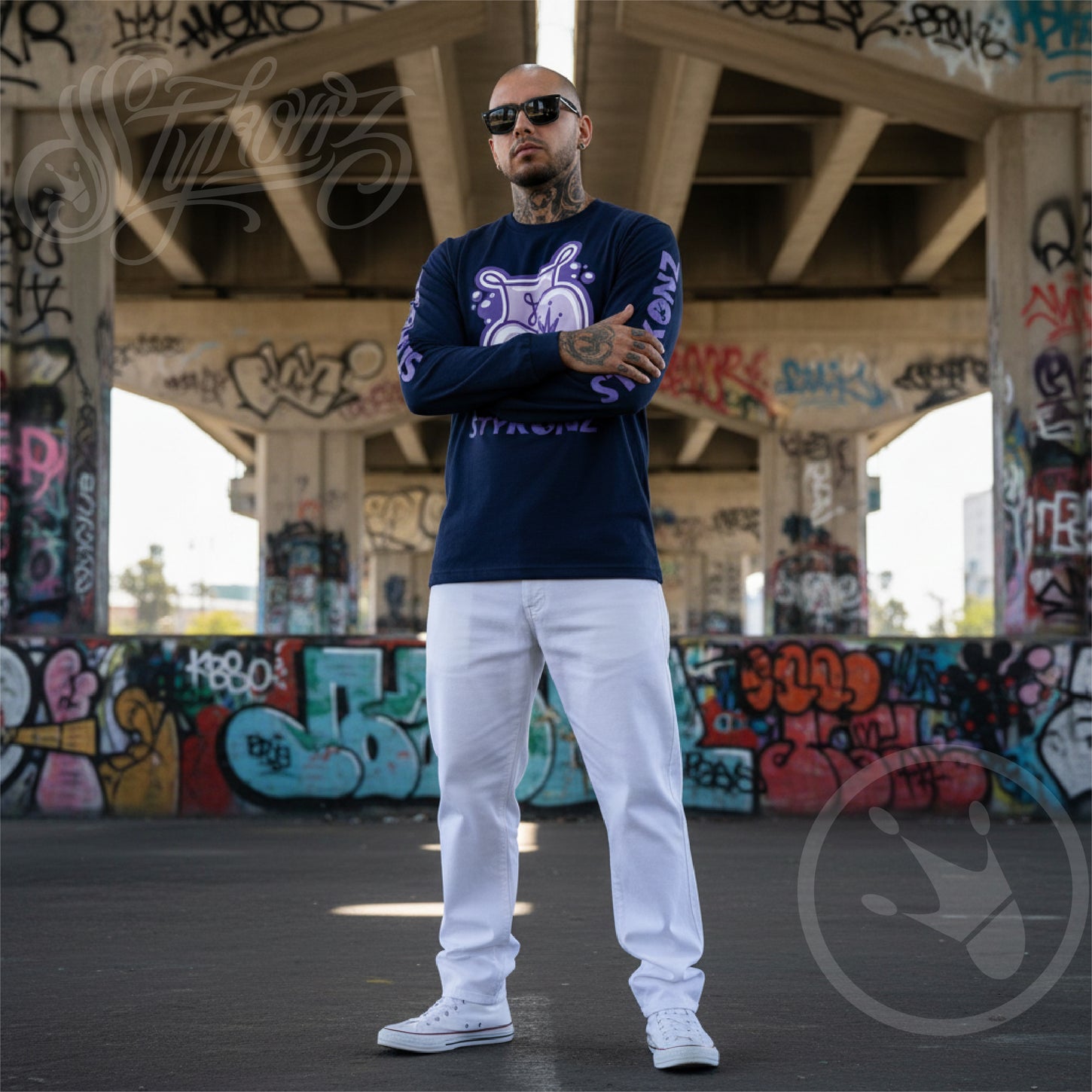 A man wearing a navy blue sweatshirt with Stykonz Throw-up, white pants, and white sneakers is standing in front of graffiti-covered walls.